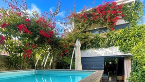 Ein Schwimmbecken mit Holzterrasse, umgeben von üppiger Vegetation und roten Blüten.