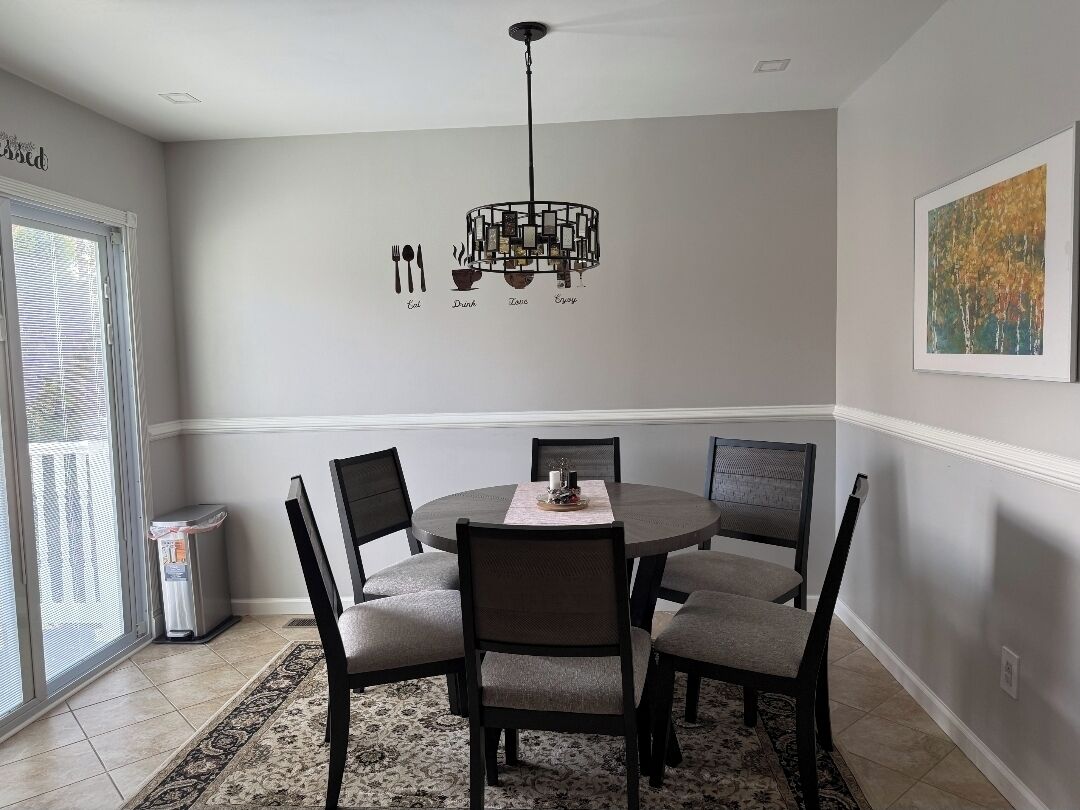 A dining area with a round table, six chairs, a chandelier, and a painting on the wall.
