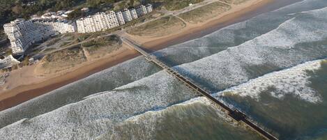 Vue aérienne d’une zone côtière avec une longue jetée s’étendant dans la mer, entourée d’un hôtel en bord de mer composé de plusieurs bâtiments, et d’une plage en arrière-plan.