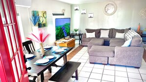 Una sala de estar con un sofá gris, una mesa de comedor con platos y vasos, un televisor y arte mural que representa paisajes urbanos.