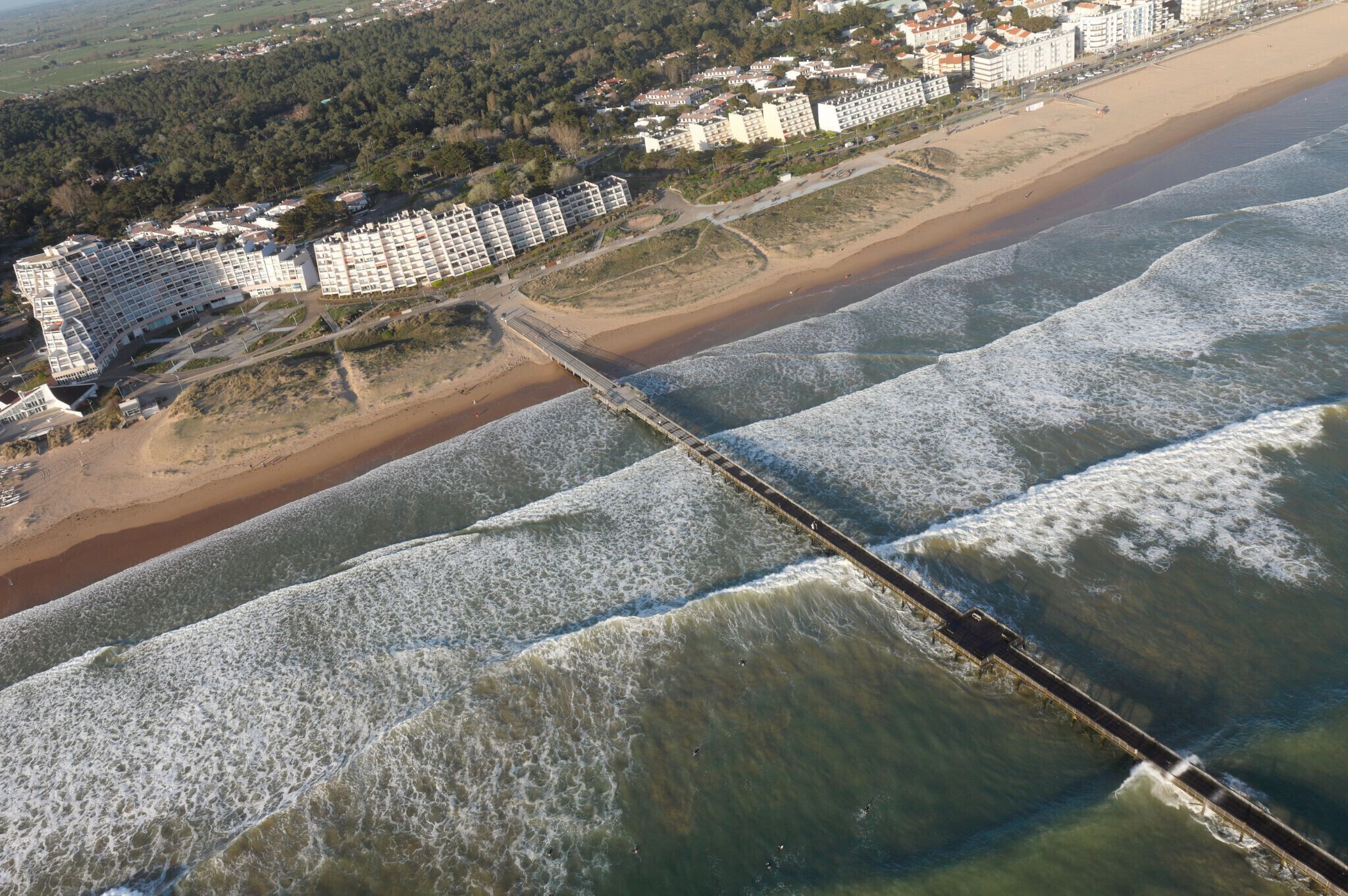 An aerial view of a coastal area with a long pier extending into the sea, surrounded by a beachfront hotel with multiple buildings and a beach in the background.