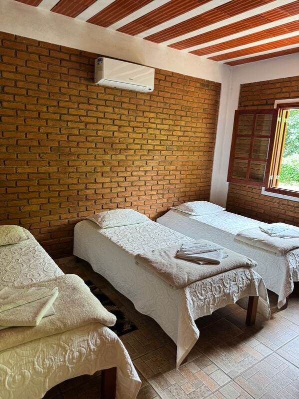 A room with two beds, a brick wall, a window with a view, and a ceiling with wooden beams. - Hotel Fazenda Bavária (Pouso Alto)
