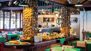 A buffet setup with various food items and a stone pillar. Tables are covered with green tablecloths, and the room has a rustic wooden ceiling. - Hotel Fazenda Bavária (Pouso Alto)