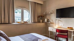 A hotel room with a bed, a desk, a chair, a TV, a window with curtains, and a view of the outside.