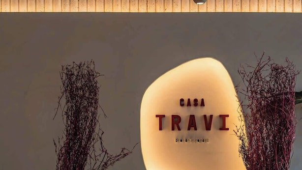 A wall-mounted sign with the text 'CASA TRAVI BOUTIQUE' in a modern font, flanked by two white vases with dried branches, set against a tiled wall with a wooden slat ceiling.