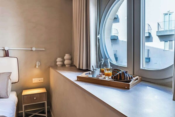 A hotel room with a bed, bedside table, window with curtains, and a breakfast tray on the windowsill.