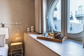 A hotel room with a bed, a bedside table with a lamp, a window with a view of buildings, and a breakfast tray with pastries and drinks.
