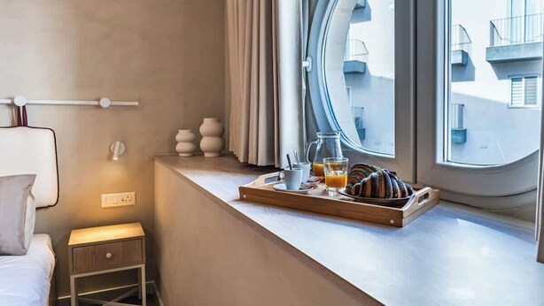 Habitació d'hotel amb llit, taula de nit, finestra amb cortines i safata de petit esmorzar al davant de la finestra.