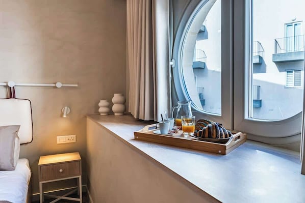 A hotel room with a bed, bedside table, window with curtains, and a breakfast tray on the windowsill.