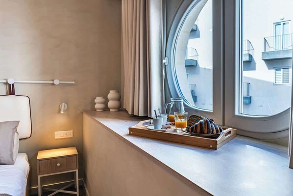 A hotel room with a bed, a bedside table with a lamp, a window with a view of buildings, and a breakfast tray with pastries and drinks.