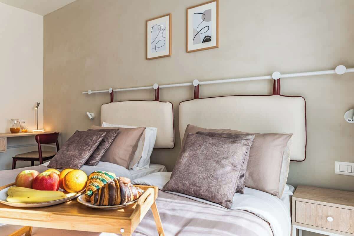 A hotel room with two beds, a breakfast tray with fruits and pastries, a bedside table, and a wall-mounted towel rack.