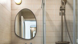 A modern bathroom with a vanity, a round mirror, a shower with a handheld showerhead, and a wall-mounted light fixture. - Travi Boutique By Holi 18 (Saint Julian's)