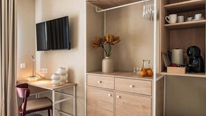 A hotel room with a desk, chair, TV, wooden cabinet, and shelves with cups and a coffee maker. - Travi Boutique By Holi 18 (Saint Julian's)