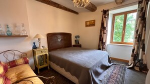 Un dormitorio con cabecera de madera, cama con manta gris, mesita de noche con lámpara, ventana con cortinas y vistas al exterior con vegetación.