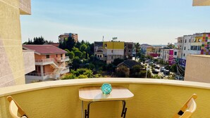 A balcony with two wooden chairs and a small table, overlooking a cityscape with various buildings and greenery. - Divjaka Apartments City Center - 212 (Divjakë)