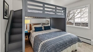 Una habitación con literas, una escalera, una mesita de noche con lámpara y una ventana con vista a árboles.