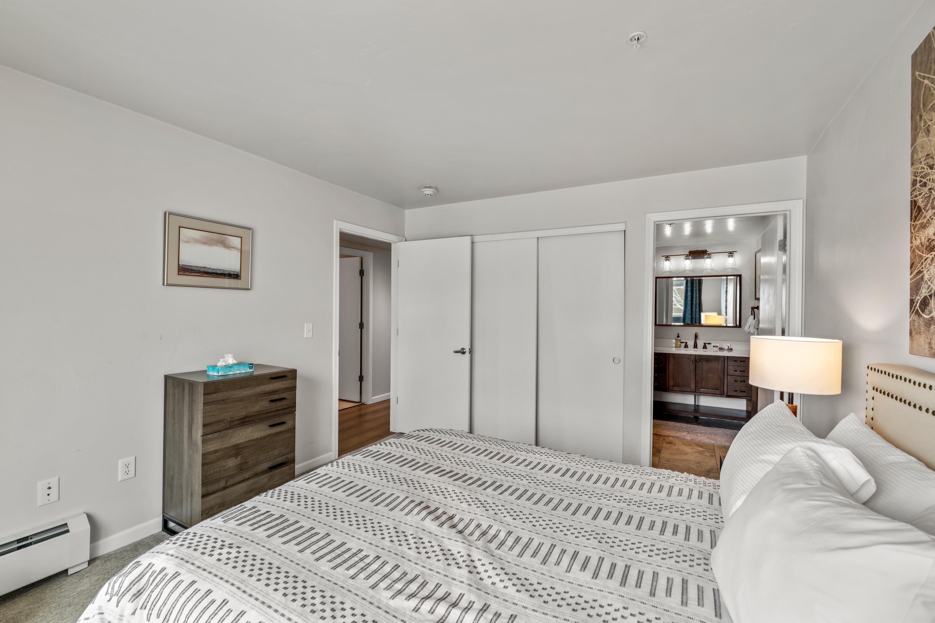 A bedroom with a bed, a dresser, a mirror, and a bathroom visible through an open door.
