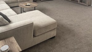 Ein Wohnzimmer mit einem Ecksofa, einem kleinen runden Couchtisch, einem grauen Sessel und einem an der Wand befestigten Fernseher.