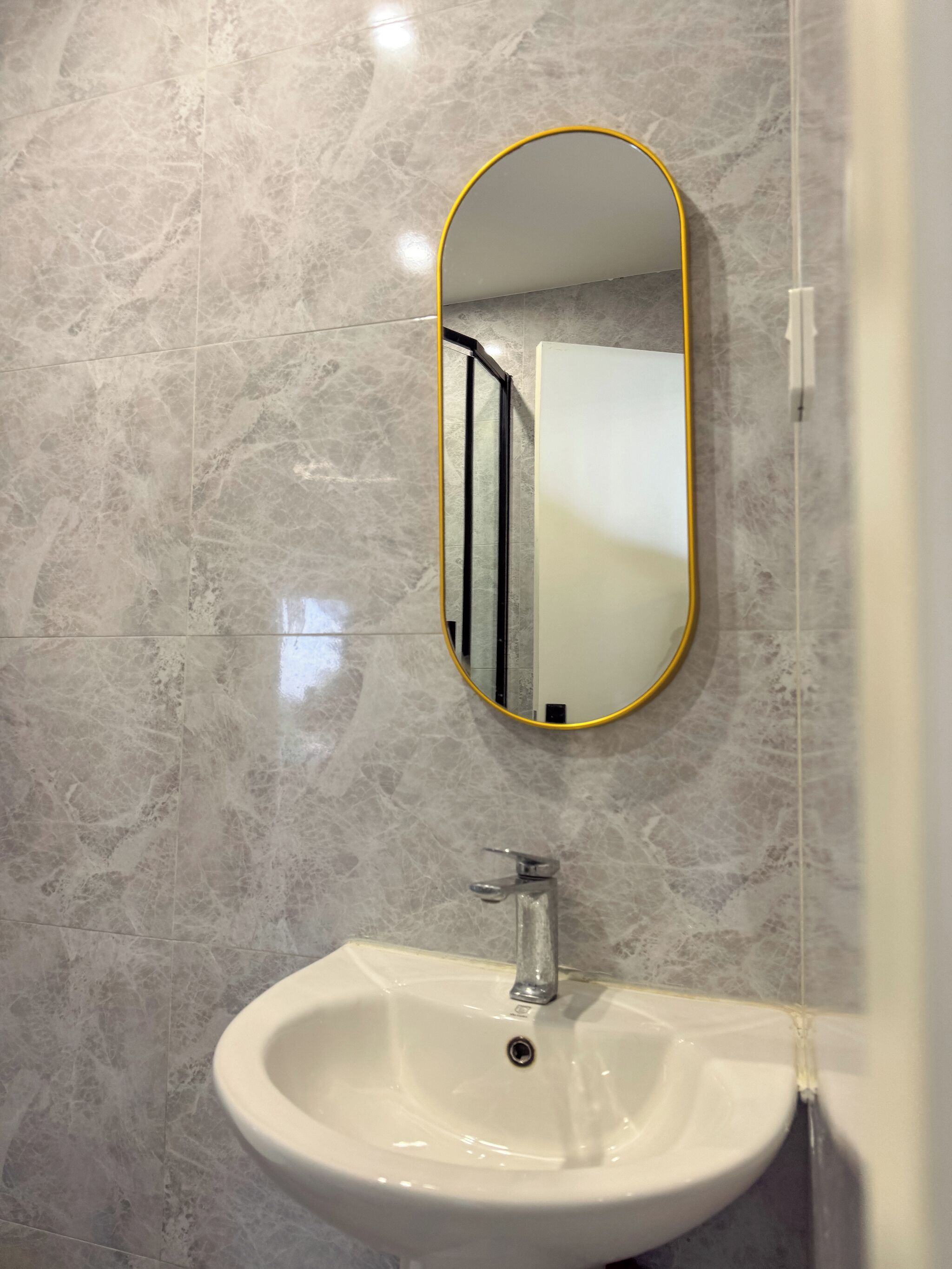 Un baño con lavabo blanco, espejo con marco amarillo y paredes de mármol.