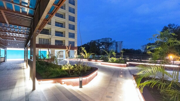 Un espacio al aire libre con un paseo techado, jardineras con follaje y un edificio al fondo.