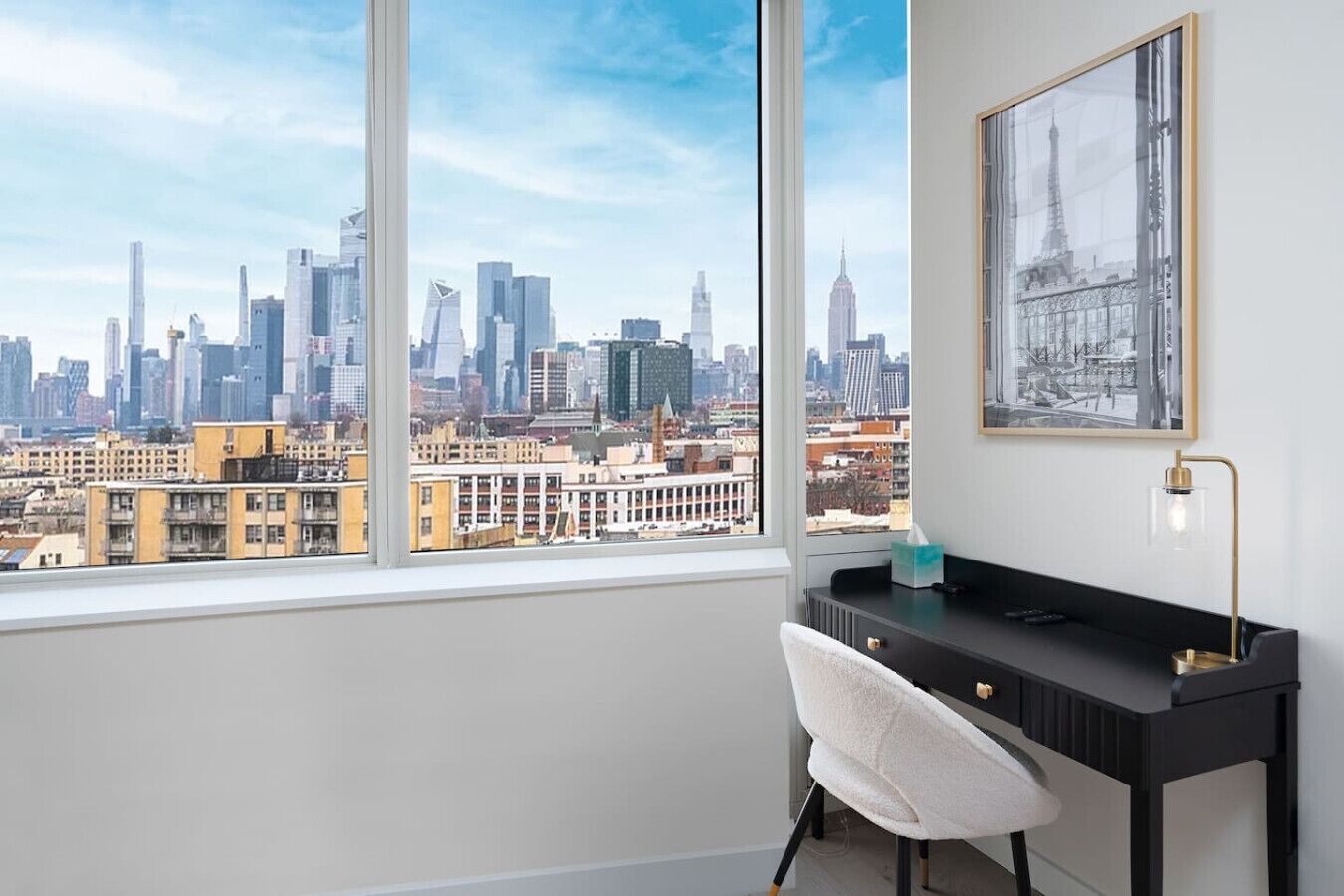 Una habitación moderna con un ventanal grande que ofrece vistas al horizonte de la ciudad, un escritorio negro, una silla blanca y un cuadro en la pared.