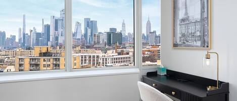 Una habitación moderna con un ventanal grande que ofrece vistas al horizonte de la ciudad, un escritorio negro, una silla blanca y un cuadro en la pared.