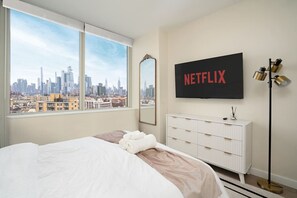 A hotel room with a large bed, a white dresser, a mirror, a wall-mounted TV displaying 'NETFLIX', and a city view.