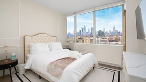 Een hotelkamer met een groot bed, een nachtkastje, een lamp en uitzicht op een stadshemel door een groot raam.