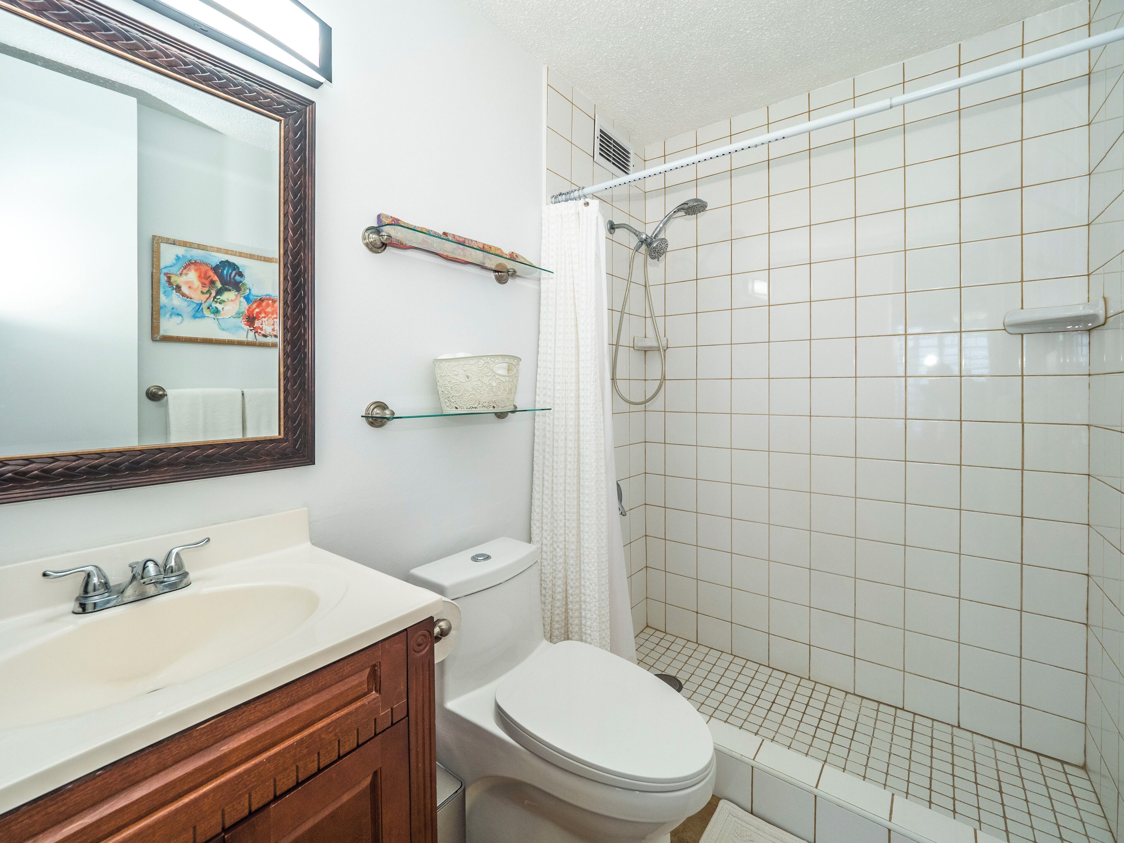 Un baño con un lavamanos blanco, tocador de madera, inodoro, cabina de ducha con mampara de vidrio y espejo con marco decorativo.