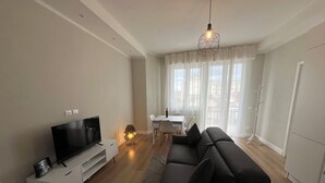 A modern living room with a flat-screen TV, a black sofa, a dining table with chairs, and a large window with curtains.