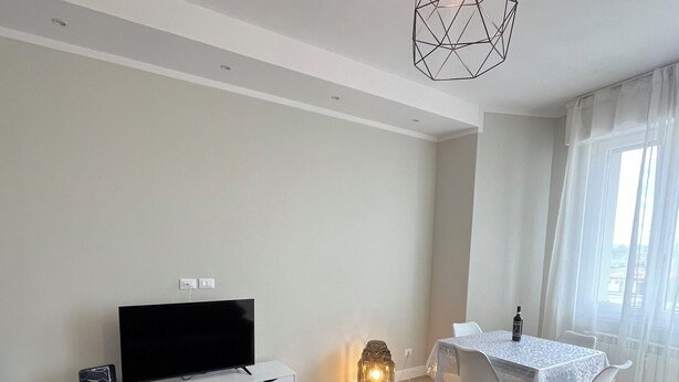 A modern living room with a grey sofa, a flat-screen TV, a white entertainment unit, a dining table with chairs, and a hanging light fixture.