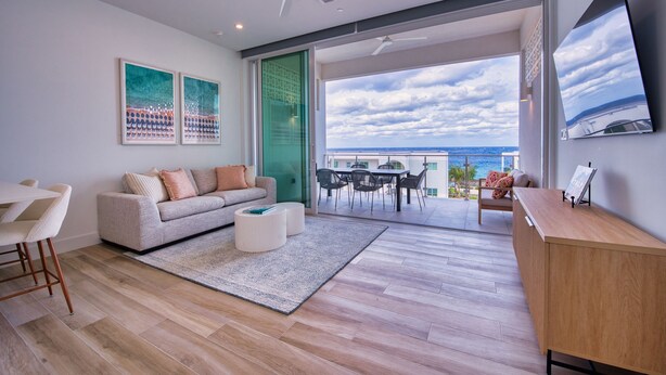Una sala de estar moderna con sofá, mesa de centro y comedor. Un ventanal ofrece vista al océano.