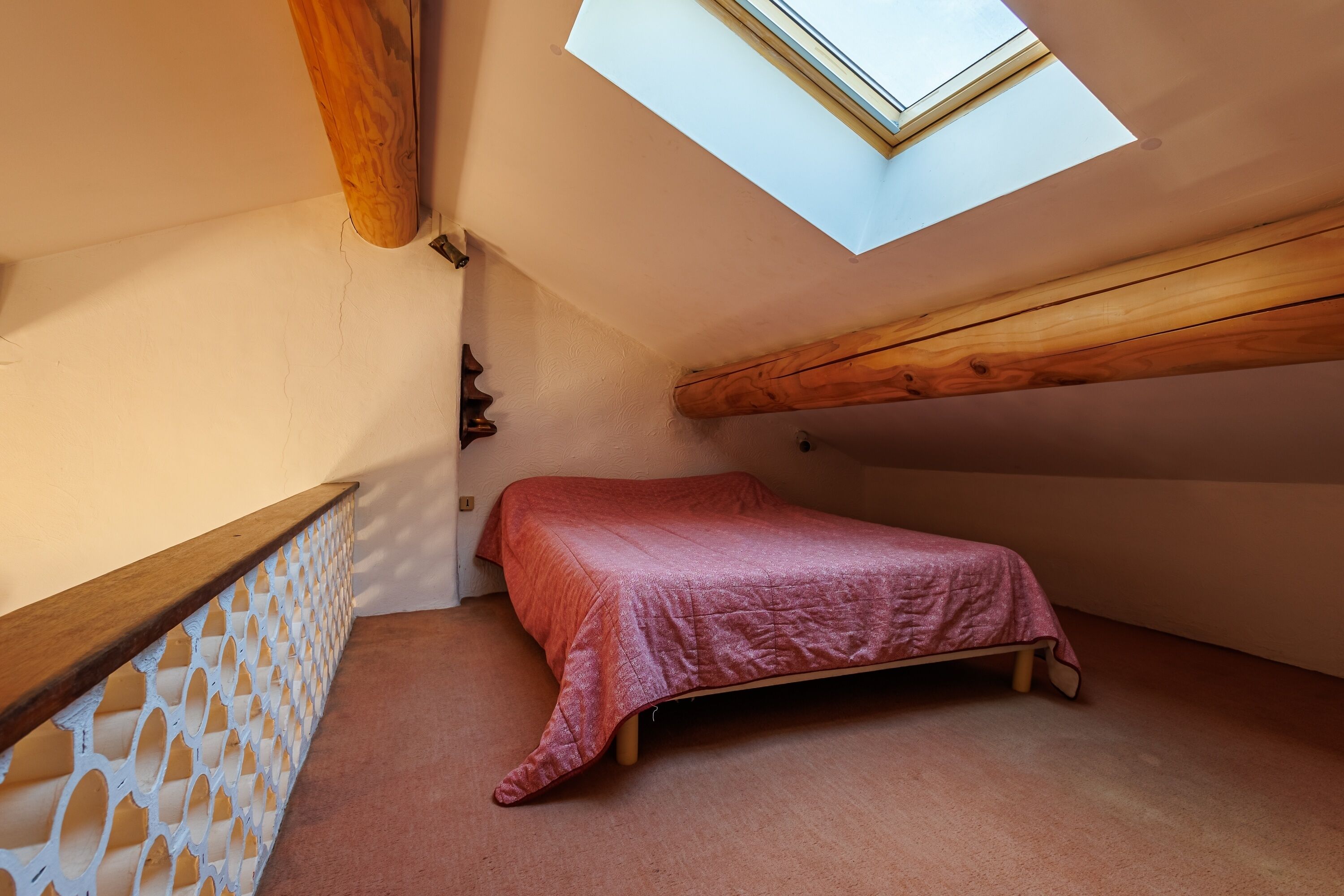 Une petite pièce avec un plafond en pente et une lucarne, une poutre en bois, un lit recouvert d’une couverture rose et un mur blanc orné de motifs.