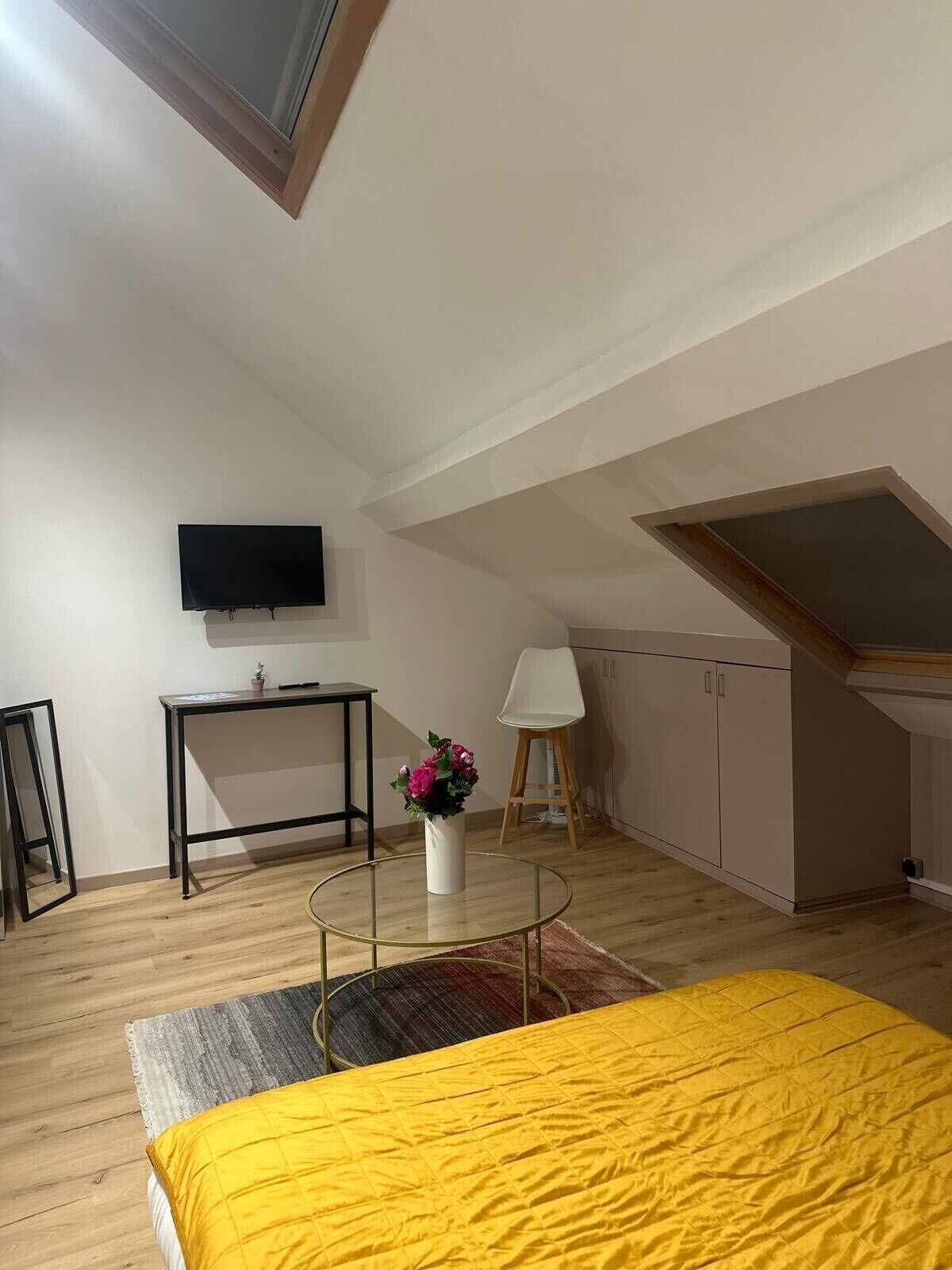 A small, well-lit room with a bed, a desk, a chair, a TV, and a window.