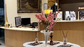 A hotel reception area with a desk, a vase of pink flowers, a framed painting, and a round table with a blue carpet.