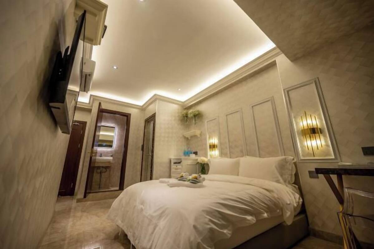 A modern bedroom with a large bed, a flat-screen TV mounted on the wall, and a small bathroom visible through an open door.