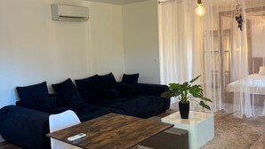 Un salon moderne avec un canapé d’angle bleu foncé, une table basse en bois et un fauteuil blanc. On aperçoit une plante en pot sur la table basse, ainsi qu’un luminaire suspendu au plafond.