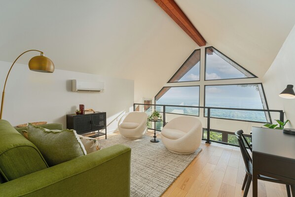 A modern living room with a green sofa, two white armchairs, a wooden dining table, and a large window offering a view of the landscape.