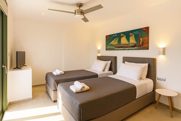 Habitación de hotel con dos camas, un ventilador de techo, un cuadro de un velero, un televisor sobre un mueble bajo y una mesita de noche.