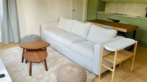 Un salon comprenant un canapé, une petite table en bois, un pouf rond et un coin cuisine avec des meubles verts et des luminaires suspendus.