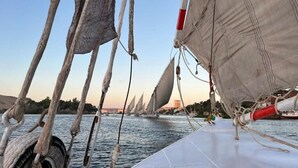 En segelbåt med flera master och segel, förtöjd nära en strandpromenad med byggnader i bakgrunden.