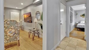 Un pasillo con un sillón floral, una mesita con lámpara, un cuadro en la pared y una cama en la habitación contigua.