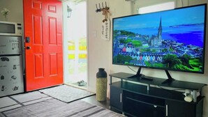 A living room with a large flat-screen TV, a red door, a kitchen area with appliances, and a decorative vase. - Craven Cottage Studio at Hillside with Wifi, AC & 4K Smart TV (El Paso)