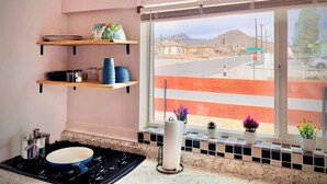 A kitchen with a window view, shelves with kitchenware, and a stove. - Craven Cottage Studio at Hillside with Wifi, AC & 4K Smart TV (El Paso)