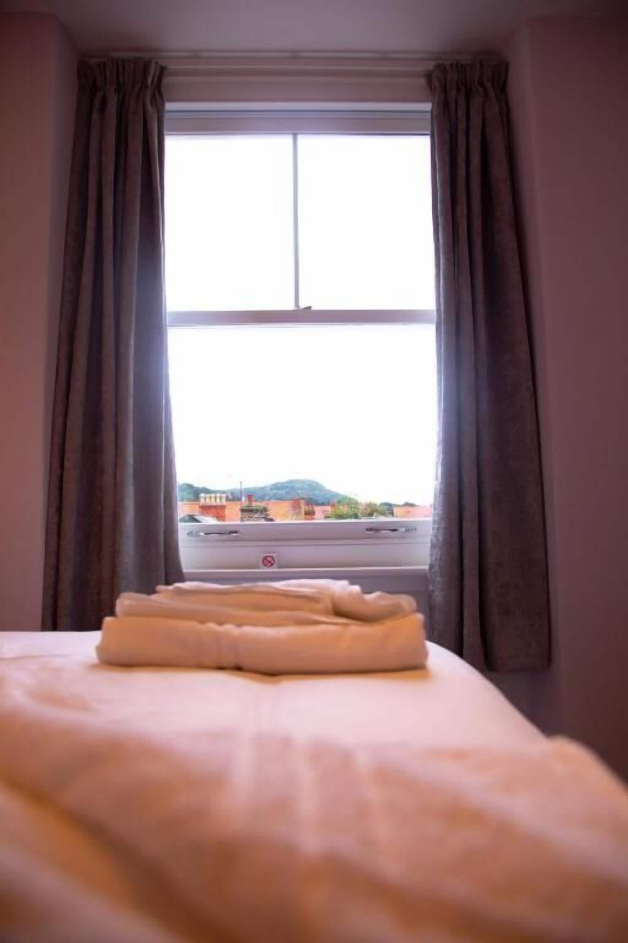 Une chambre avec un lit, une fenêtre avec des rideaux et une vue sur les bâtiments et les collines environnants.