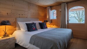 A wooden cabin bedroom with a bed, pillows, a nightstand, and a window with curtains. - Chalet San Marco - mountain luxury stay for 6 - OVO Network (La Clusaz)