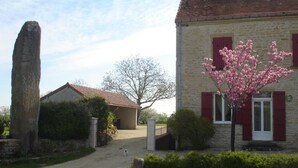 Un edificio de piedra con puerta y contraventanas rojas, un pilar de piedra grande y un árbol con flores rosadas.