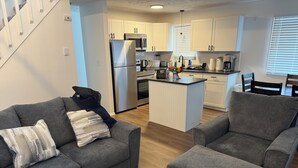 A modern kitchen with white cabinets, a stainless steel refrigerator, and a microwave. There is a gray sofa with a decorative pillow and a wooden coffee table with a floral arrangement.
