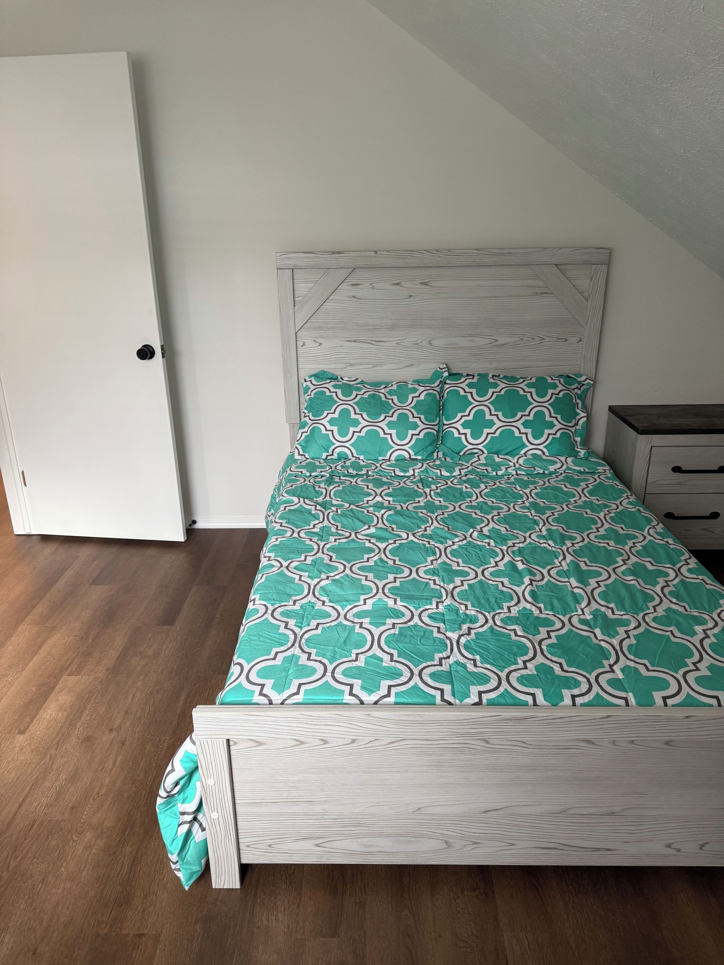 A bedroom with a wooden bed frame, a patterned bedspread, a nightstand, and a white door.