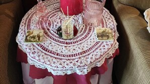 Una mesa con mantel de encaje, un florero rojo con flores, una vela y dos adornos decorativos.